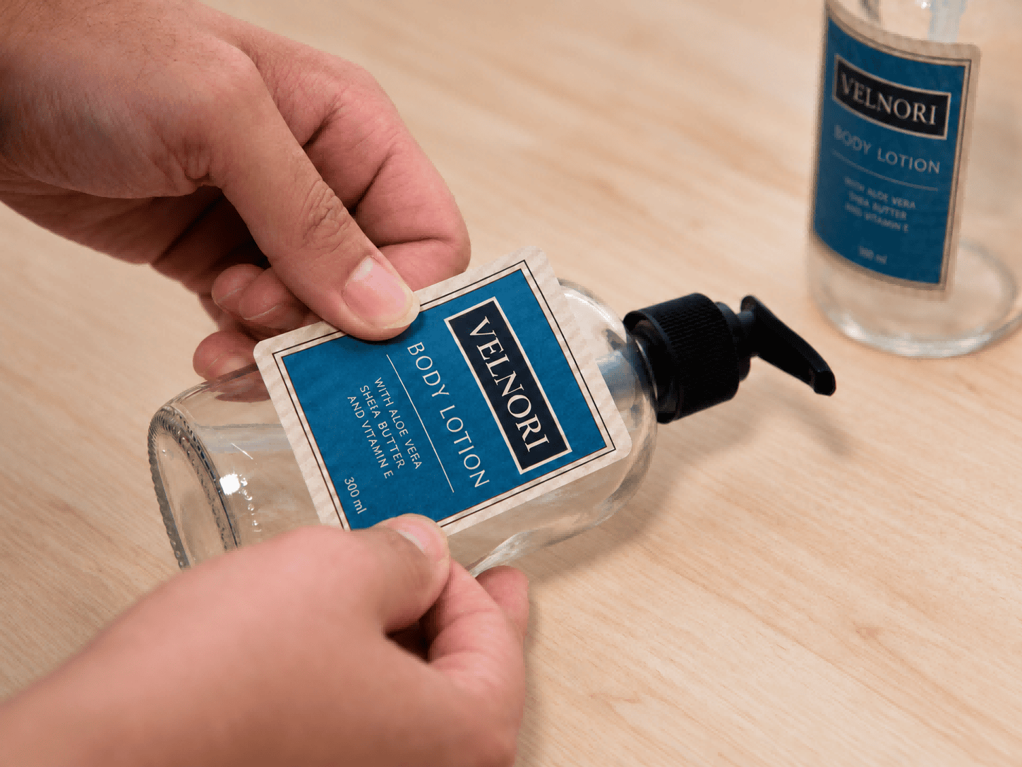 Hands pressing a blue body lotion label onto a clear bottle with a black pump