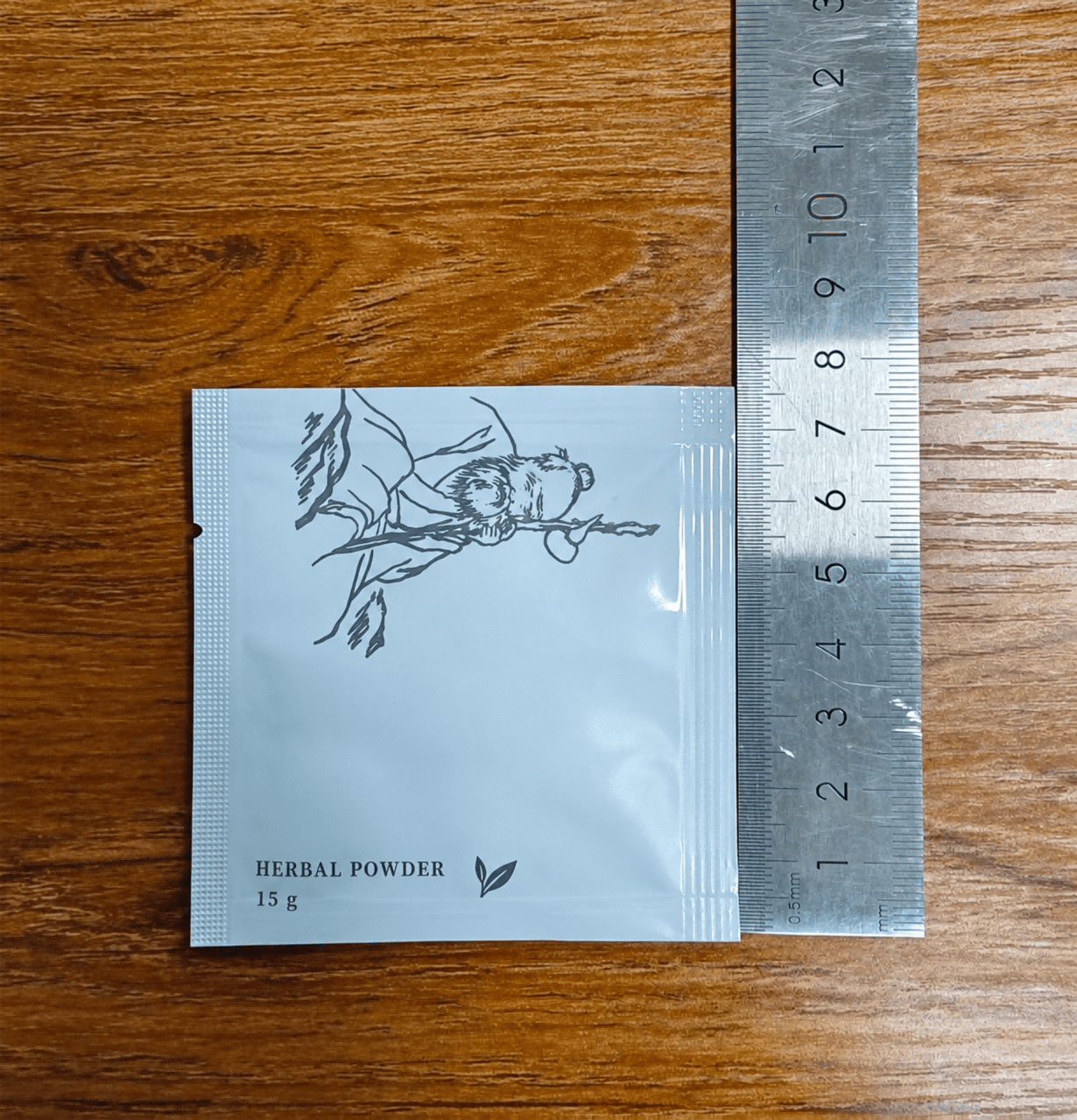 Herbal powder sachet with textured heat seals and a side tear notch beside a metal ruler on a wooden surface