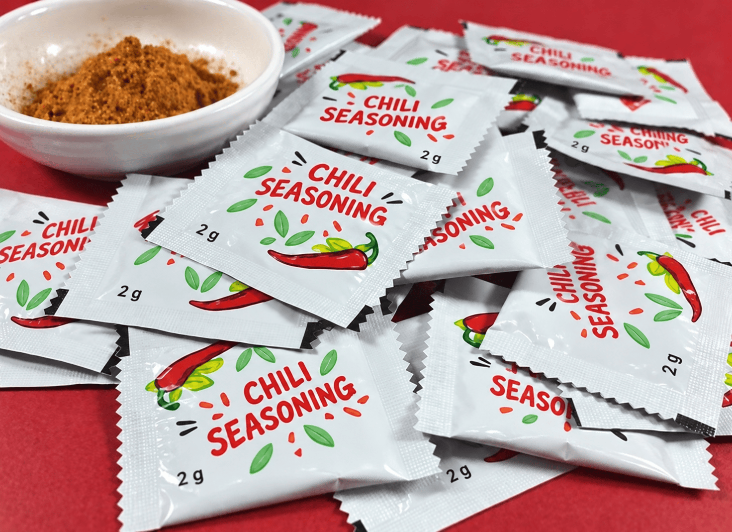 White chili seasoning sachets arranged beside a bowl of seasoning powder on a red surface