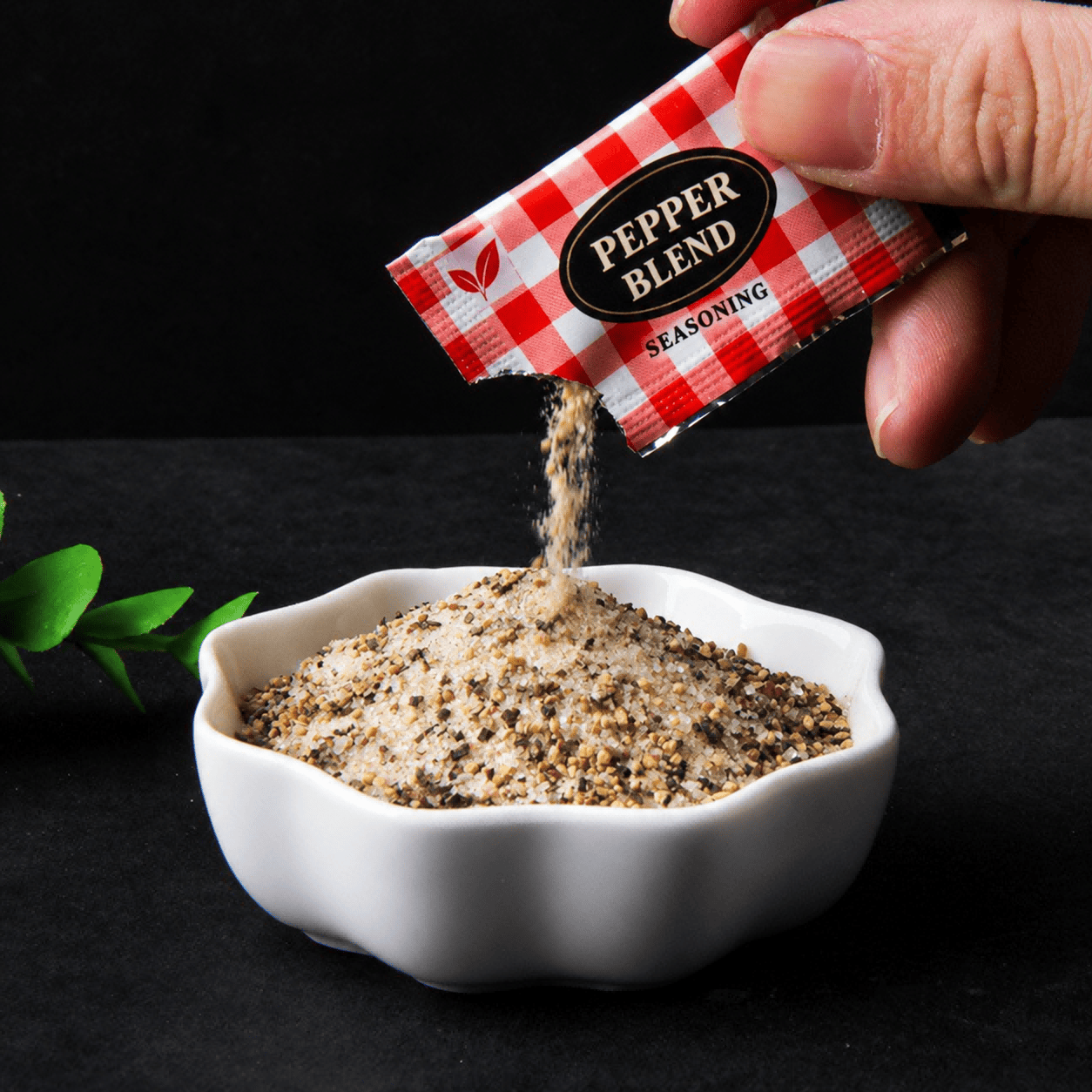 A hand pouring a coarse seasoning blend from a red check-pattern sachet into a white bowl