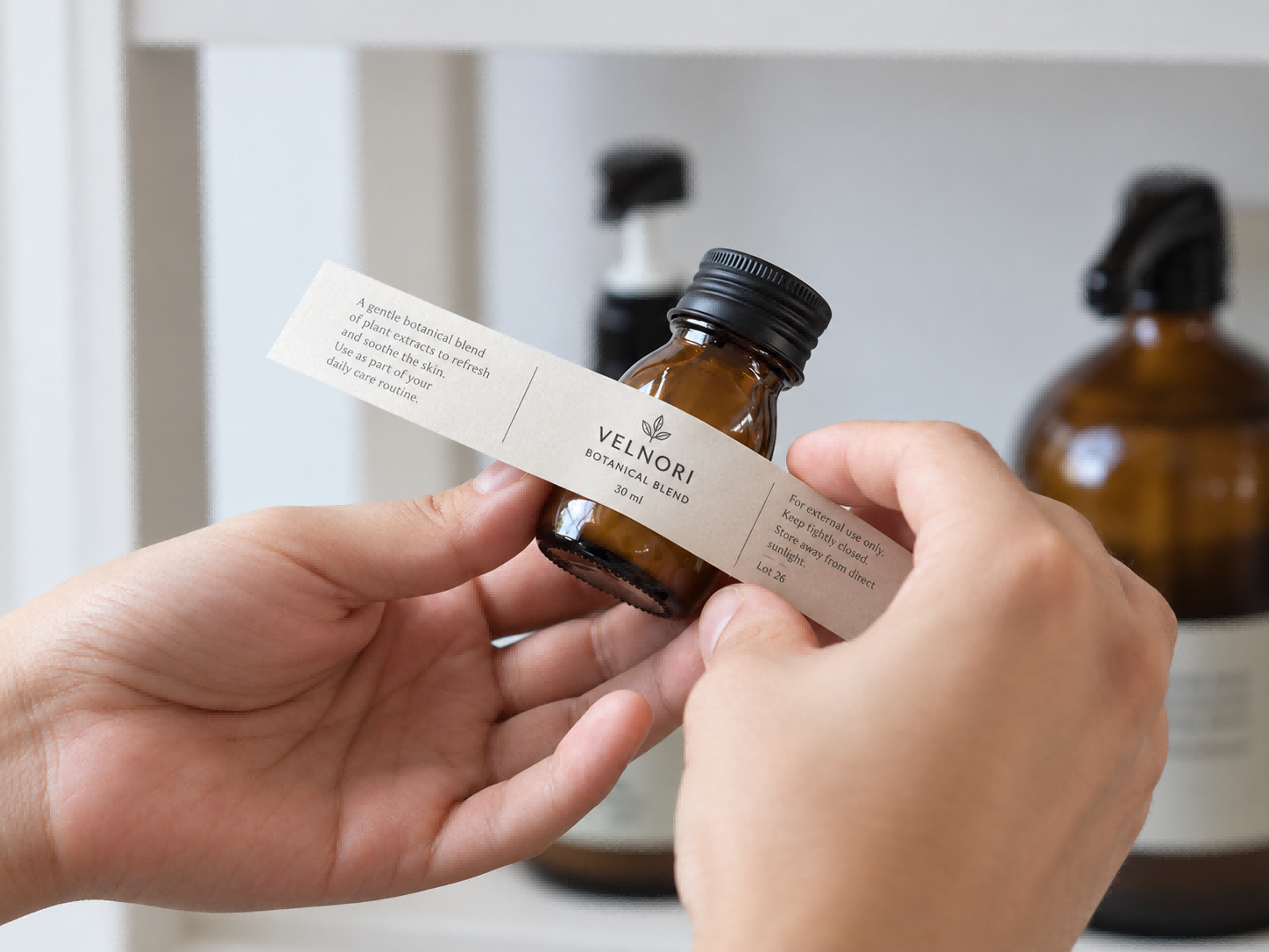 Hands aligning a narrow cream wraparound label around a small amber glass bottle